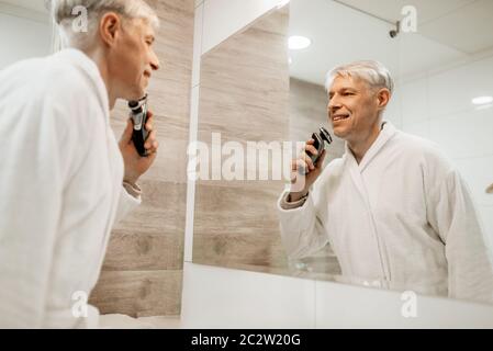 Allegro uomo adulto in accappatoio rade presso lo specchio del bagno. Grey-haired maschio maturo persona con rasoio elettrico Foto Stock