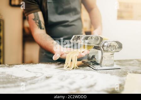 Maschio di chef nella cucina della catenaria freschi fatti in casa fettuccine in macchina per la pasta di legno sul tavolo da cucina Foto Stock