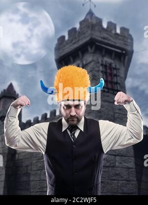 Imprenditore dispari nel casco di partito che mostra il suo strenth contro la torre antica Foto Stock