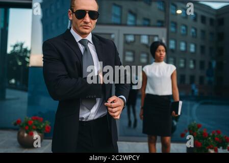 Bodyguard gravi in tuta, occhiali da sole e l'auricolare, femmina client su sfondo. La guardia di sicurezza è una professione rischiosa, professional a guardia di busine Foto Stock