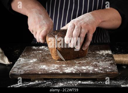 Chef in uniforme nera contiene un coltello da cucina in mano e taglio dei pezzi di pane da un marrone al forno farina di segale focaccia su una tavola di legno Foto Stock