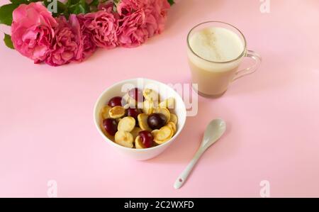 Una ciotola bianca con cereali, una piccola frittella con ciliegie e una tazza di cappuccino, un bouquet di rose su sfondo colorato. Cucina alla moda. Foto Stock