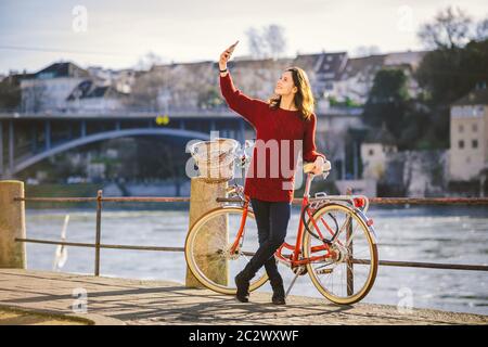 Una bella giovane donna con una bicicletta rossa retrò sta facendo una foto di se stessa nella città vecchia d'Europa sul fiume Reno emban Foto Stock