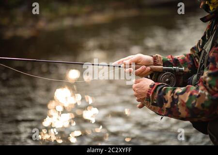 Vista ravvicinata delle mani di un pescatore a mosca che lavorano la linea e la canna da pesca mentre la pesca con la mosca su di uno splendido fiume di montagna per la trota iridea Foto Stock