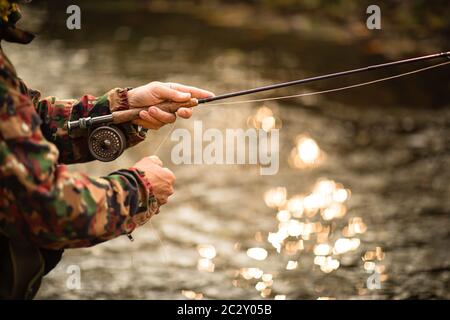 Vista ravvicinata delle mani di un pescatore a mosca che lavorano la linea e la canna da pesca mentre la pesca con la mosca su di uno splendido fiume di montagna per la trota iridea Foto Stock