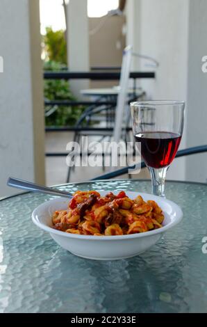 Primo piano di una ciotola bianca di pasta ripiena (ravioli) con salsa di pomodoro e funghi con un bicchiere di vino rosso su un tavolo da balcone esterno Foto Stock