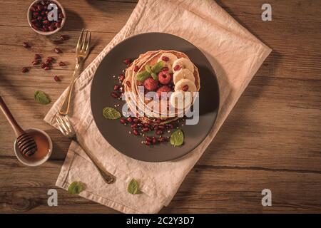 Pancake con lamponi, fette di banana, semi di melograno e miele su tavola d'annata in legno. Foto Stock