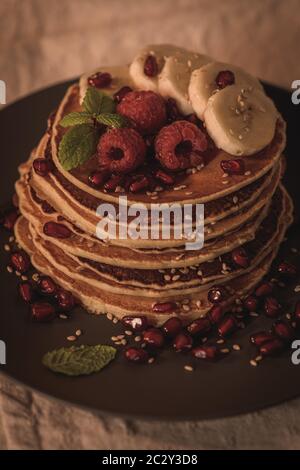 Pancake con lamponi, fette di banana, semi di melograno e miele su tavola d'annata in legno. Foto Stock