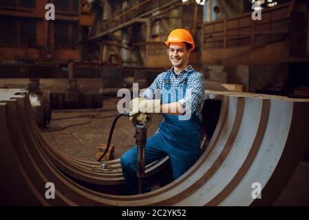 Lavoratore maschile in uniforme e casco rimuove la bilancia da pezzi di metallo in fabbrica. Industria metallurgica, produzione industriale di prodotti siderurgici Foto Stock