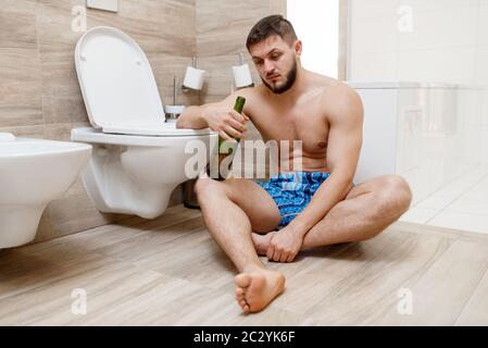 Uomo di sbornia con bottiglia di vino seduto sul pavimento vicino al gabinetto in bagno. Uomo di hungover, mal di testa e depressione, mattina cattivo Foto Stock
