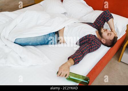 L'uomo ubriaco dorme a letto con una bottiglia di vino. Uomo con alcol hangover, mal di testa e depressione, cattivo mattino Foto Stock