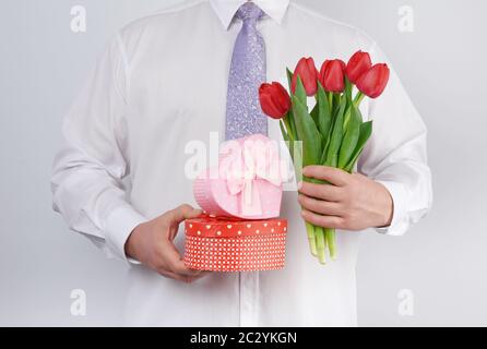 uomo adulto in camicia bianca e cravatta lilla con bouquet di tulipani rossi con foglie verdi e scatola regalo su sfondo bianco, concetto per bir felice Foto Stock