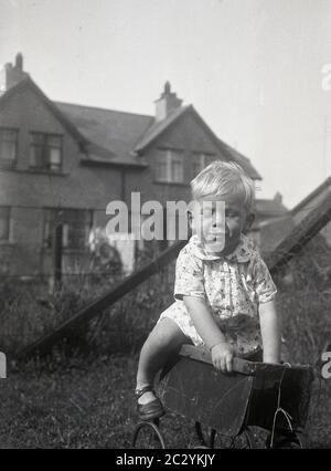 circa anni '30, storico, diurno e un ragazzino che gioca all'aperto in un giardino, arrampicandosi su un vecchio giocattolo di un bambino con ruote in metallo o su un pram di bambola dell'epoca, Che ha perso il suo cappuccio e maniglia, un oggetto interessante da provare e salire e il bambino mostra un buon equilibrio! Foto Stock