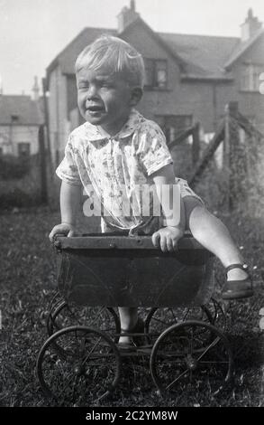 Circa1930, storico, diurno e un ragazzino che gioca all'esterno in un giardino, arrampicandosi sulla cima di un vecchio giocattolo di un bambino con ruote in metallo o di un pRAM di bambola dell'epoca, che ha perso il suo cappuccio e maniglia, un oggetto interessante da provare e salire e il bambino mostra un buon equilibrio! Foto Stock