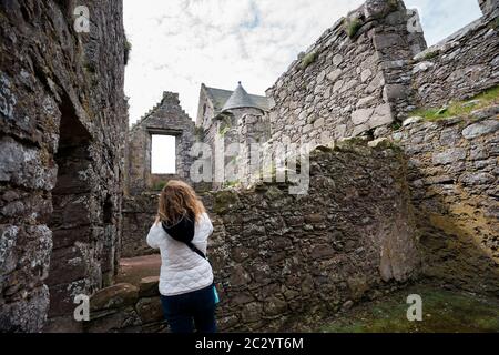 Un turista biondo esplora le mura che si sbriciolavano sotto un cielo nuvoloso nel castello in pietra rovinato di Dunnottar, Stonehaven, Scozia, Regno Unito Foto Stock