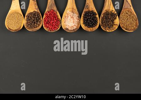 Sette cucchiai da cucina in legno d'oliva, pieni di spezie varie, si trovano in una fila su sfondo nero con spazio per le copie Foto Stock