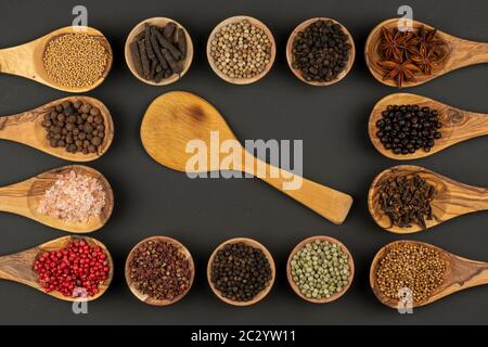 Sei piccole ciotole in legno e otto cucchiai da cucina in legno di ulivo riempiti di varie spezie e un singolo cucchiaio da cucina in legno si trovano su un grigro nero Foto Stock