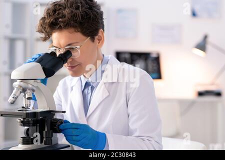 Medico scienziato con capelli ricci in camice da laboratorio e guanti in lattice utilizzando il microscopio mentre si cerca la mutazione del coronavirus in laboratorio Foto Stock