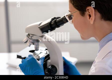 Lavoratore di laboratorio concentrato in guanti utilizzando il microscopio durante l'analisi della struttura cellulare Foto Stock