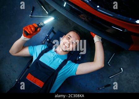 Lavoratore in uniforme giacente sotto il veicolo, stazione di servizio auto. Controllo e ispezione di automobili, diagnostica professionale e riparazione Foto Stock