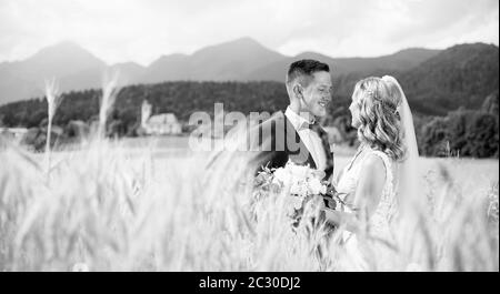 Lo sposo abbracci sposa teneramente nel campo di grano da qualche parte nella campagna slovena. Caucasico romantico felice coppia giovane per celebrare il loro matrimonio. Black n Foto Stock