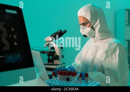 Ricercatore coronavirus focalizzato analizzando il sangue e prendendo appunti sulla malattia utilizzando il laptop in laboratorio medico Foto Stock