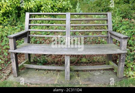 Tradizionale in legno curvato park sede sulla pavimentazione in pietra lastre circondato da piante con erba in primo piano. Foto Stock