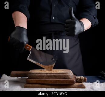 cuoco maschio in uniforme nera e guanti di lattice nero tiene un grande coltello di carne tagliente su un tagliere, un'altra mano mostra come gesto, backgrou nero Foto Stock