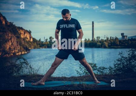 Uomo ispirato che fa asana yoga sul lago di cava. Giovane cittadino che si esercita all'esterno e si trova in posizione di yoga angolo laterale. Fitness all'aperto e balanc di vita Foto Stock
