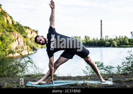 Uomo ispirato che fa asana yoga sul lago di cava. Giovane cittadino che si esercita all'esterno e si trova in posizione di yoga angolo laterale. Fitness all'aperto e balanc di vita Foto Stock