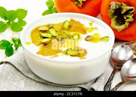 Yogurt, persimmon e dolce al miele con vaniglia, il cardamomo e i pistacchi in una ciotola su un tovagliolo, menta e versare sulla tavola di legno sfondo Foto Stock