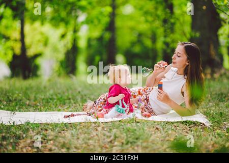 Donna sorridente giocare e sedersi su erba verde nel parco, riposo e abbraccio tenere sapone soffiatore bolla con bambina carina bambino. Felice Foto Stock