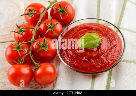 Gustoso e denso ketchup di pomodoro in una ciotola di vetro e pomodori ciliegia rossi maturi su un rametto sopra il tavolo da cucina in legno bianco. Spezie, condimenti e salse. Foto Stock