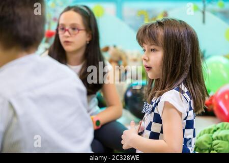 Vista laterale di un simpatico e ragazza timida guardando i suoi colleghi più anziani che interagiscono durante la riproduzione libera in aula di una moderna kindergarten Foto Stock