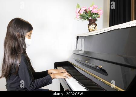 Una studentessa asiatica che indossa una maschera e suona il pianoforte a casa. L'immagine mostra l'idea durante la quarantena domestica per ridurre la diffusione della corona Foto Stock