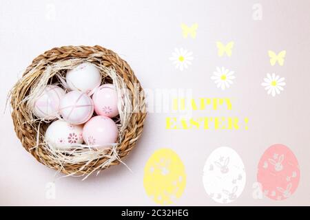 Buona cartolina di auguri di Pasqua. Vista dall'alto delle uova colorate con paglia in un cesto di vimini su uno sfondo rosa chiaro con iscrizione Happy easter e Bloss Foto Stock