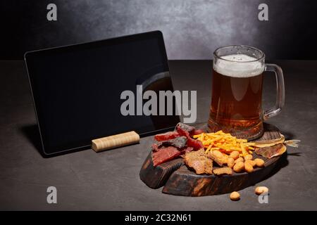 Tazza di vetro con birra, set di spuntini di vario tipo e tavoletta vuota per computer su tavolo grigio scuro su sfondo grigio. Spuntini per la birra. Foto Stock