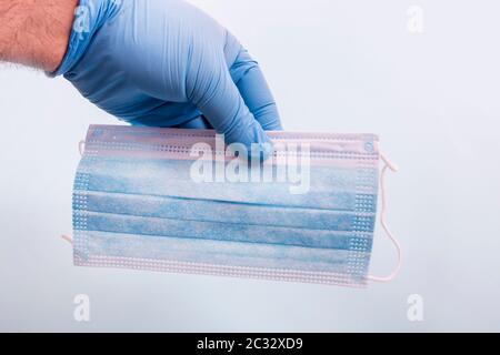 mano del medico nei guanti blu tiene la maschera facciale monouso su uno sfondo blu chiaro. Foto Stock