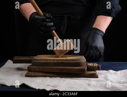 cuoco maschile in uniforme nera contiene un martello di legno per battere la carne su un tagliere vintage, sfondo nero Foto Stock