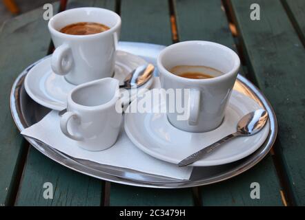 Chiudere uno argento vassoio di metallo bianco con tazze di caffè espresso su piattini con cucchiai e bricco per latte servita sul verde di legno verniciato tabella, elevato h Foto Stock