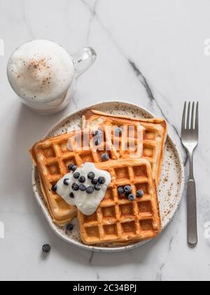 Deliziosi waffle belgi fatti in casa con yogurt greco, mirtilli e cappuccino su sfondo in marmo bianco. Colazione perfetta con spazio per fotocopie