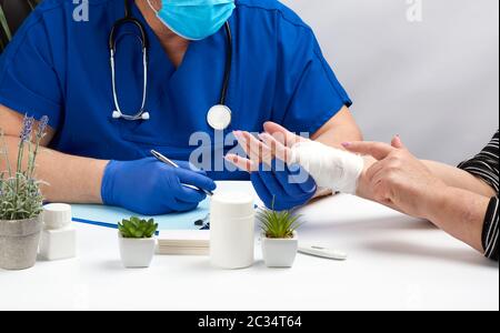 il medico traumatologo in guanti medici in lattice blu siede a un tavolo e tiene un paziente di ricezione con una ferita della mano, il posto di lavoro del medico Foto Stock