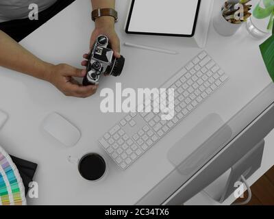 Foto a soffitto di un fotografo maschile che tiene la fotocamera sopra la scrivania bianca dell'ufficio con tablet, computer e altri materiali di consumo Foto Stock