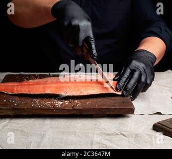 Lo chef vestiti di nero e nero Guanti in lattice tagli freschi di filetto di salmone a pezzetti, tasto scuro Foto Stock