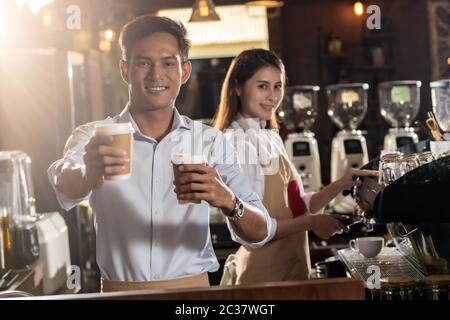 Ritratto di asian barista tenere take away tazza di caffè e che serve al cliente per cafe con altri barista lavora in background Foto Stock