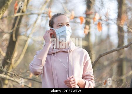 Ritratto della donna sportiva caucasica che indossa la maschera facciale di protezione medica mentre cammina nel parco, rilassandosi e ascoltando la musica. Corona virus, o Covid Foto Stock