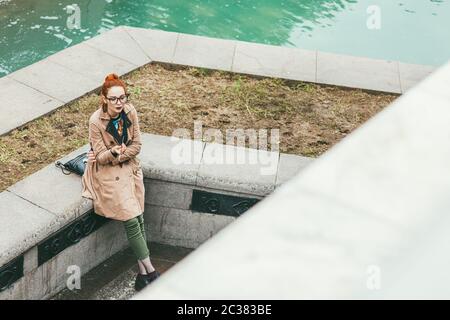 Mosca, Russia - 7 LUGLIO 2017. Una giovane donna hipster pensante in un impermeabile beige e pantaloni corti verdi e bicchieri si siede su un cu Foto Stock