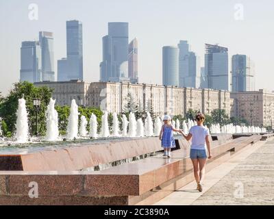 Una bambina cammina lungo il bordo della fontana in una giornata estiva soleggiata con la mano con la madre sullo sfondo di grattacieli moderni Foto Stock