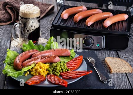 Salsicce fritte su un piatto e una griglia elettrica con un bicchiere di birra scura. Il concetto di cibo tradizionale. Foto Stock