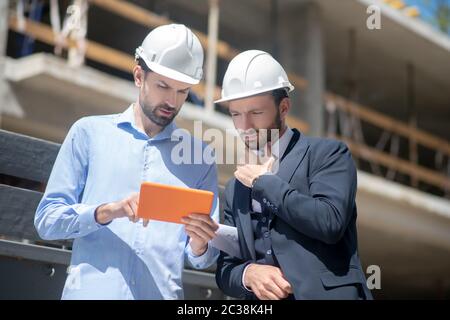 Cantiere. Caposquadra che mostra qualcosa sul suo tablet al supervisore dell'edificio, che tocca il mento Foto Stock